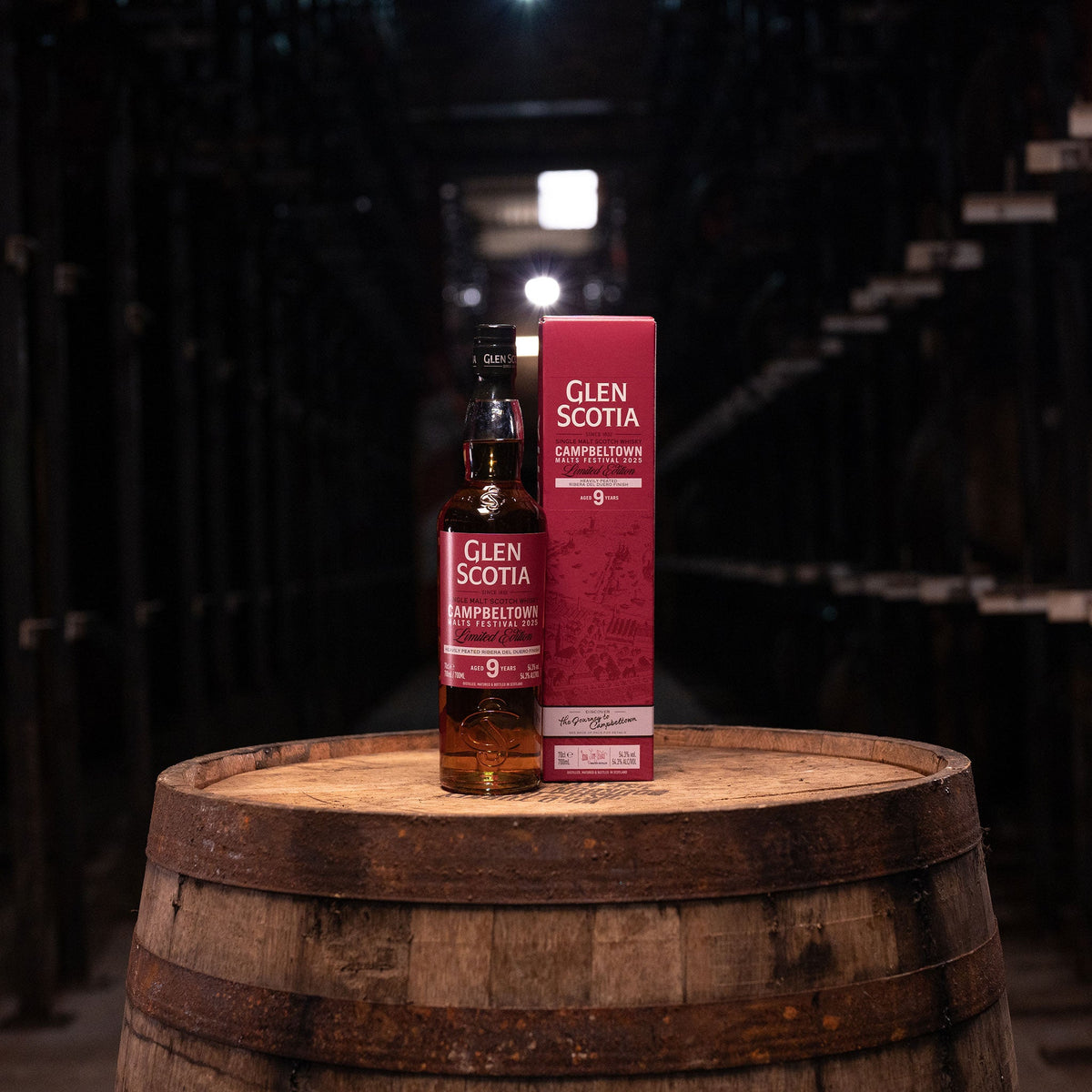 A bottle of Glen Scotia Campbeltown Malts Festival Edition 2025 rests on a wooden barrel next to its box. Finished in Ribera del Duero wine casks, it's set in a dimly lit cellar with rows of wooden casks, highlighting its unique character.