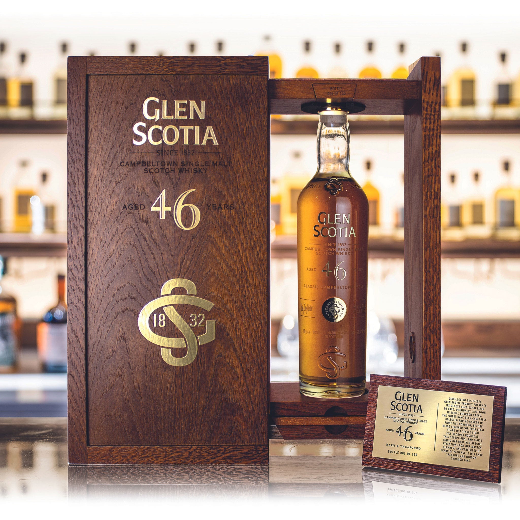 A bottle of Glen Scotia 46 Year Old Whisky, matured in a first fill bourbon cask, is displayed in a wooden case. The case has "Glen Scotia," "46 years," and "1832" engraved on it. A plaque with details about the whisky is placed in front of the bottle, with shelves of additional bottles visible in the background.
