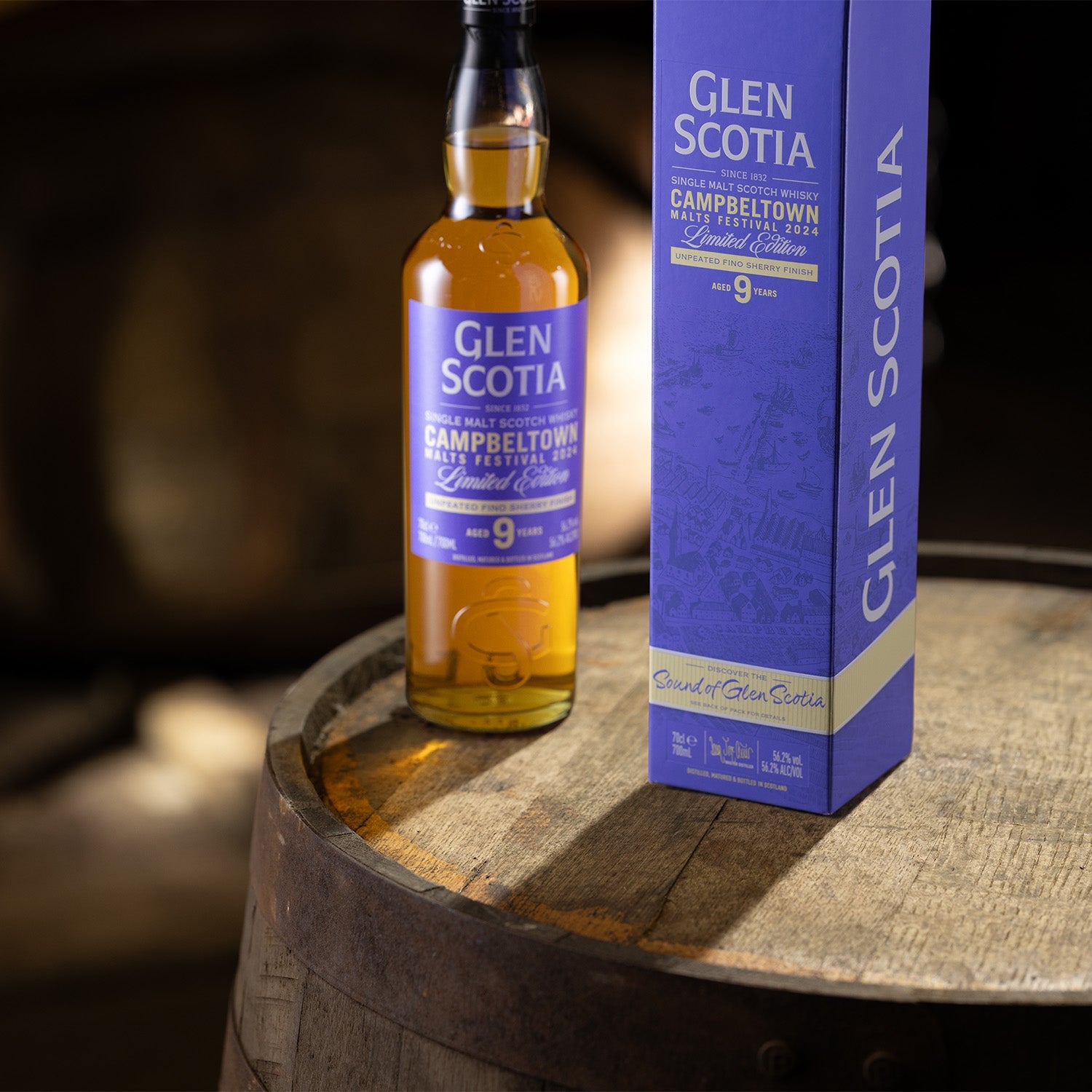 A bottle of Glen Scotia's Campbeltown Malts Festival Edition 2024 Scotch Whisky sits on a wooden barrel beside its purple and white box packaging. The label indicates it's a limited edition release—perfect for whisky gifts. The background is dimly lit.