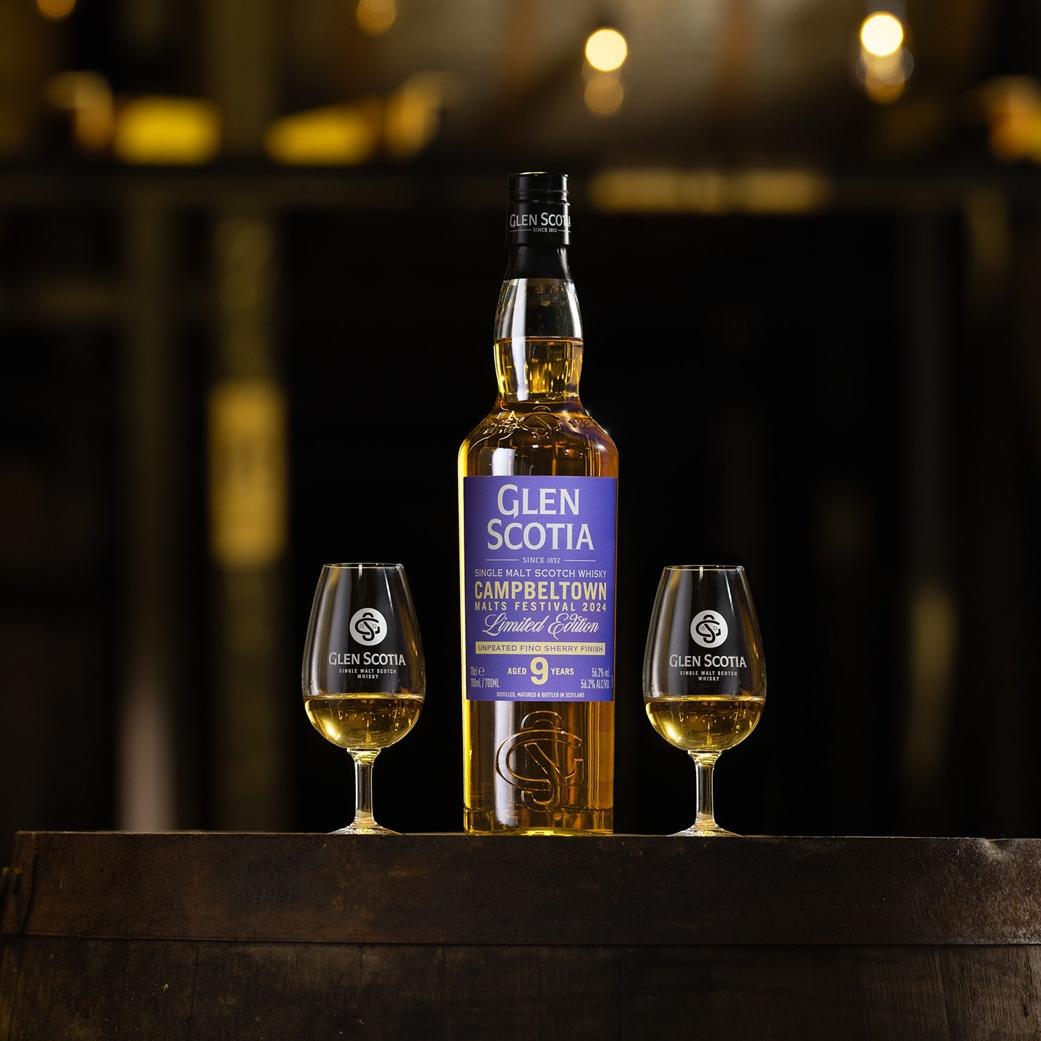 A bottle of Glen Scotia's Campbeltown Malts Festival Edition 2024 single malt scotch whisky is displayed on a table, flanked by two glasses filled with whisky. The bottle has a prominent label and is centrally positioned, ideal as a whisky gift, with a warm, ambient background.