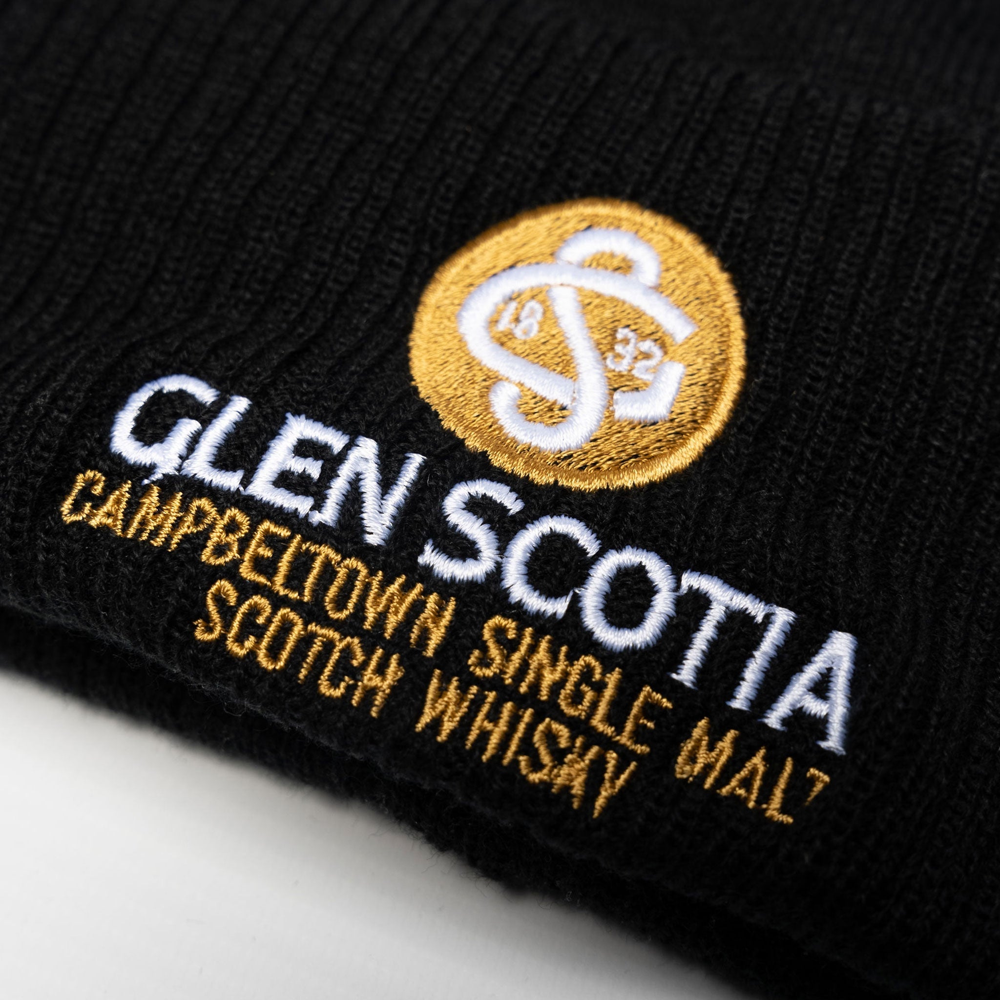 A close-up of the Glen Scotia Beanie Bobble Hat showcases white and yellow embroidery. The text "GLEN SCOTIA CAMPBELTOWN SINGLE MALT SCOTCH WHISKY" encircles the Glen Scotia logo, featuring interlocking letters "GS" and the year "1832." This cold-weather accessory from Glen Scotia masterfully combines style with heritage.