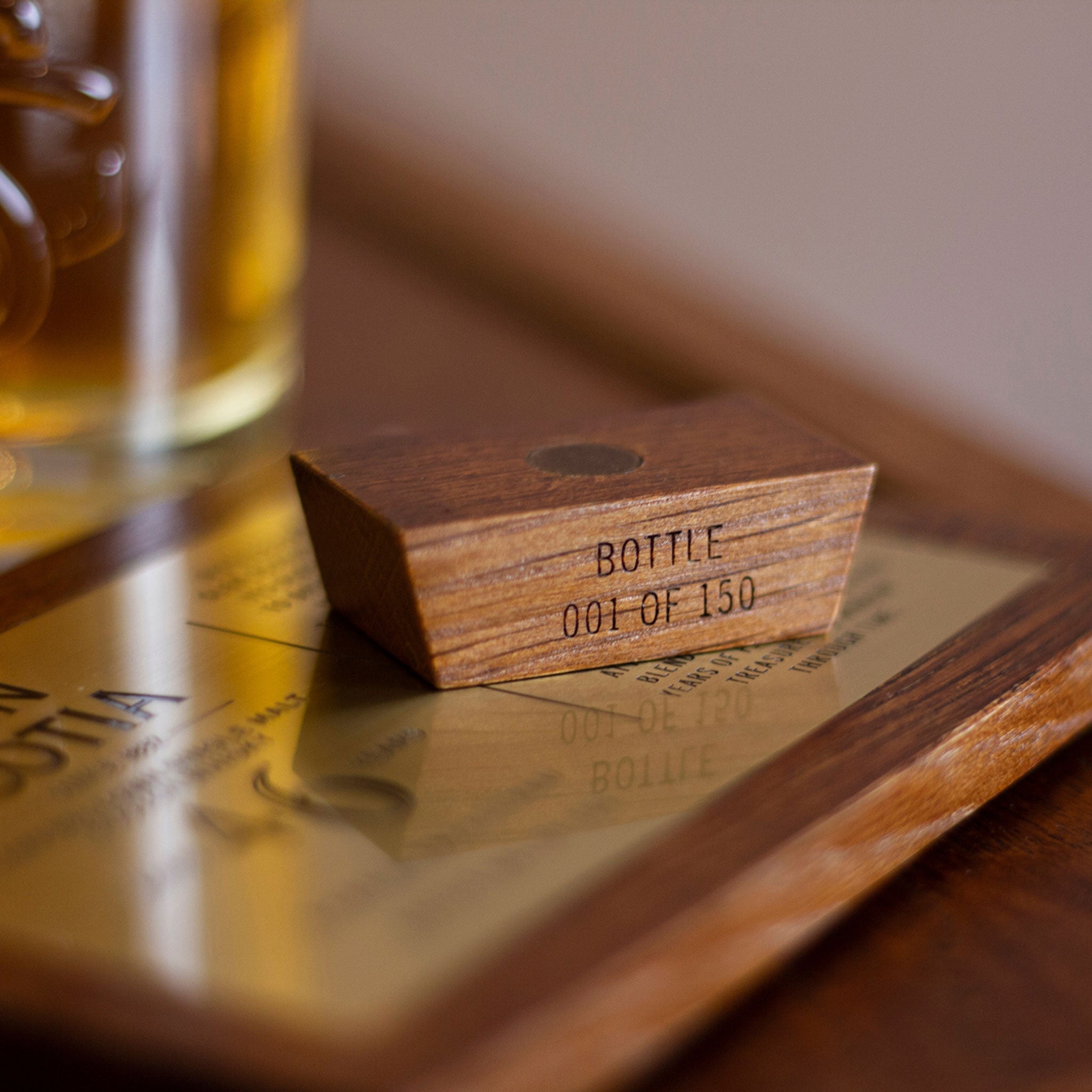 Here's a revised version of the sentence incorporating the given product data:

A close-up of a small wooden block with the text "BOTTLE 001 OF 150" engraved on it rests on a golden plaque beside a glass containing what appears to be Glen Scotia's exquisite 46 Year Old Whisky. This image beautifully captures the essence of Campbeltown climate and first fill bourbon aging.