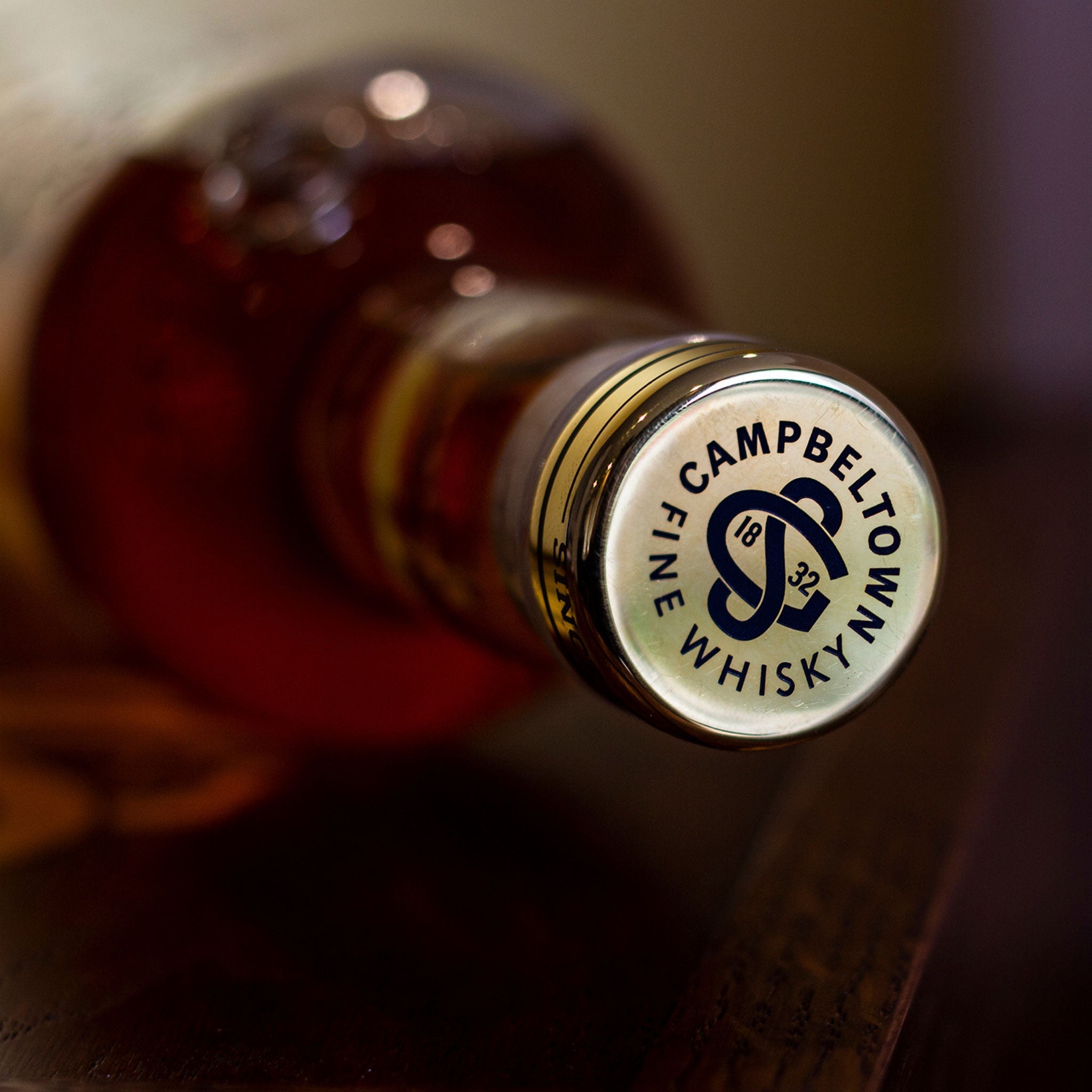 Close-up image of a whisky bottle's neck and cap lying horizontally. The cap features the label "Glen Scotia Campbeltown Fine Whisky" with a logo in the center. The bottle contains amber-colored 46 Year Old Whisky, matured in first fill bourbon casks, and the background is blurred.