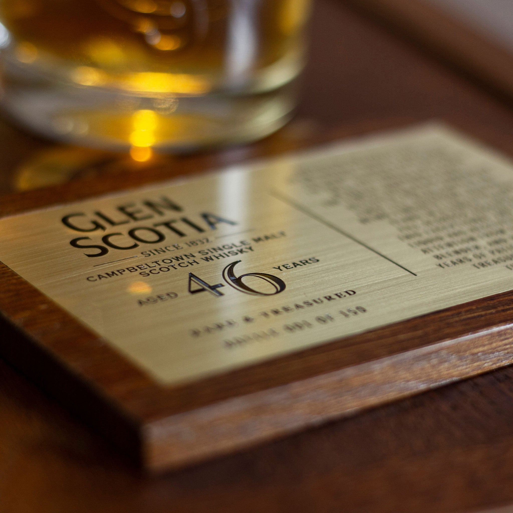 Close-up of a brass plaque on a wooden base, commemorating a 46 Year Old Whisky by Glen Scotia. Part of the plaque's text is out of focus, while a glass of whisky, aged in first-fill bourbon barrels and influenced by the Campbeltown climate, is visible in the background.