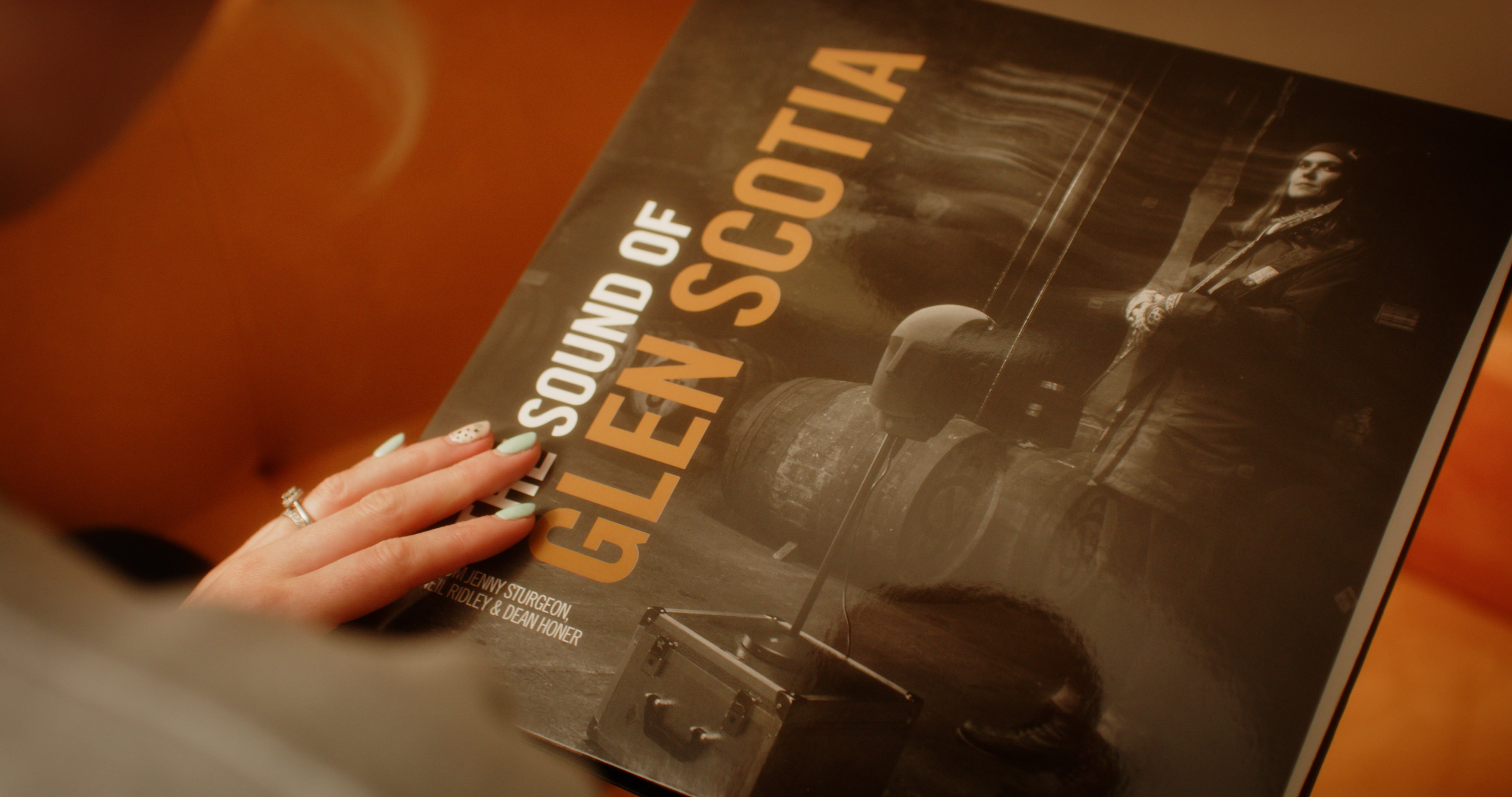 A person holds a vinyl record with the title "THE SOUND OF GLEN SCOTIA" from Glen Scotia written on the cover. The cover features someone in a kilt and traditional Scottish attire, standing next to whisky barrels in Campbeltown.