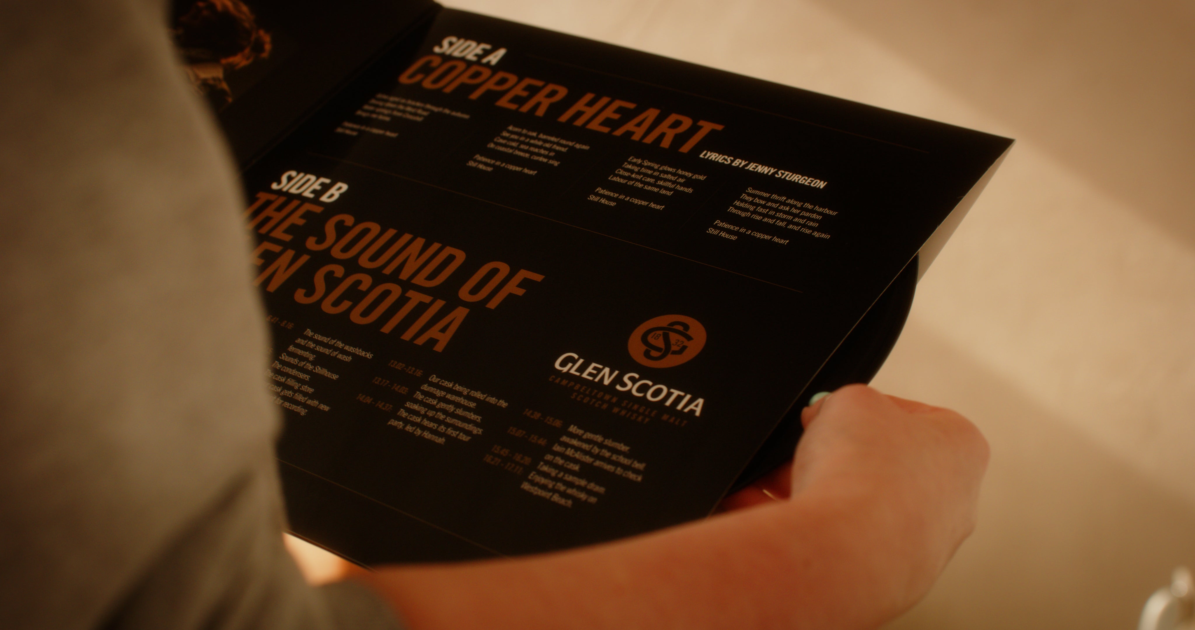 A person holds "The Sound of Glen Scotia," a black booklet from the brand Glen Scotia. It features two titled sections: "Side A: Copper Heart" and "Side B: The Sound of Glen Scotia." Each section lists events or items with descriptions. The iconic Glen Scotia logo, synonymous with Campbeltown whisky, is visible in the lower half of the right page.