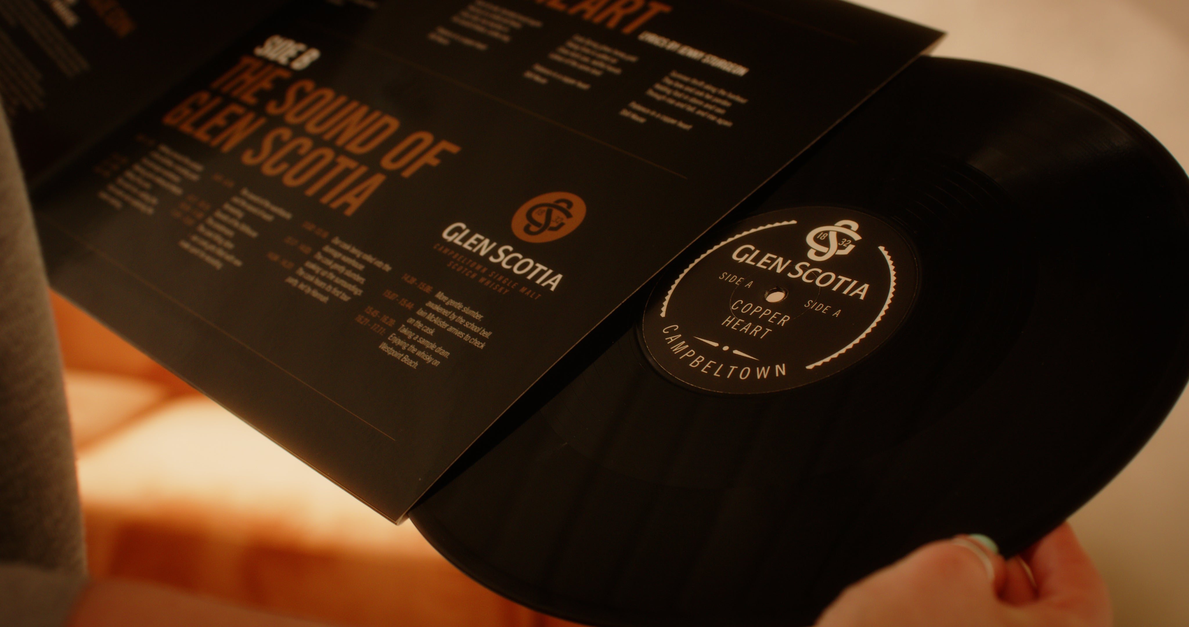 Close-up of a person holding a vinyl record partially pulled out from its sleeve. The sleeve features a design with text about "The Sound of Glen Scotia," and the label on the record reads "Glen Scotia." The warm-toned, blurred background evokes the rich heritage of Campbeltown whisky.